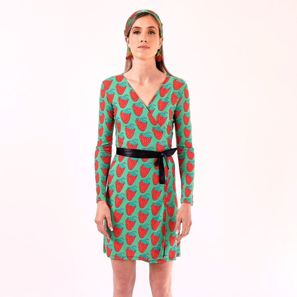 Front studio view of a model wearing Quirk’s green Tastes Like Strawberry wrap dress with red hand-drawn fruit print and a black waist tie.