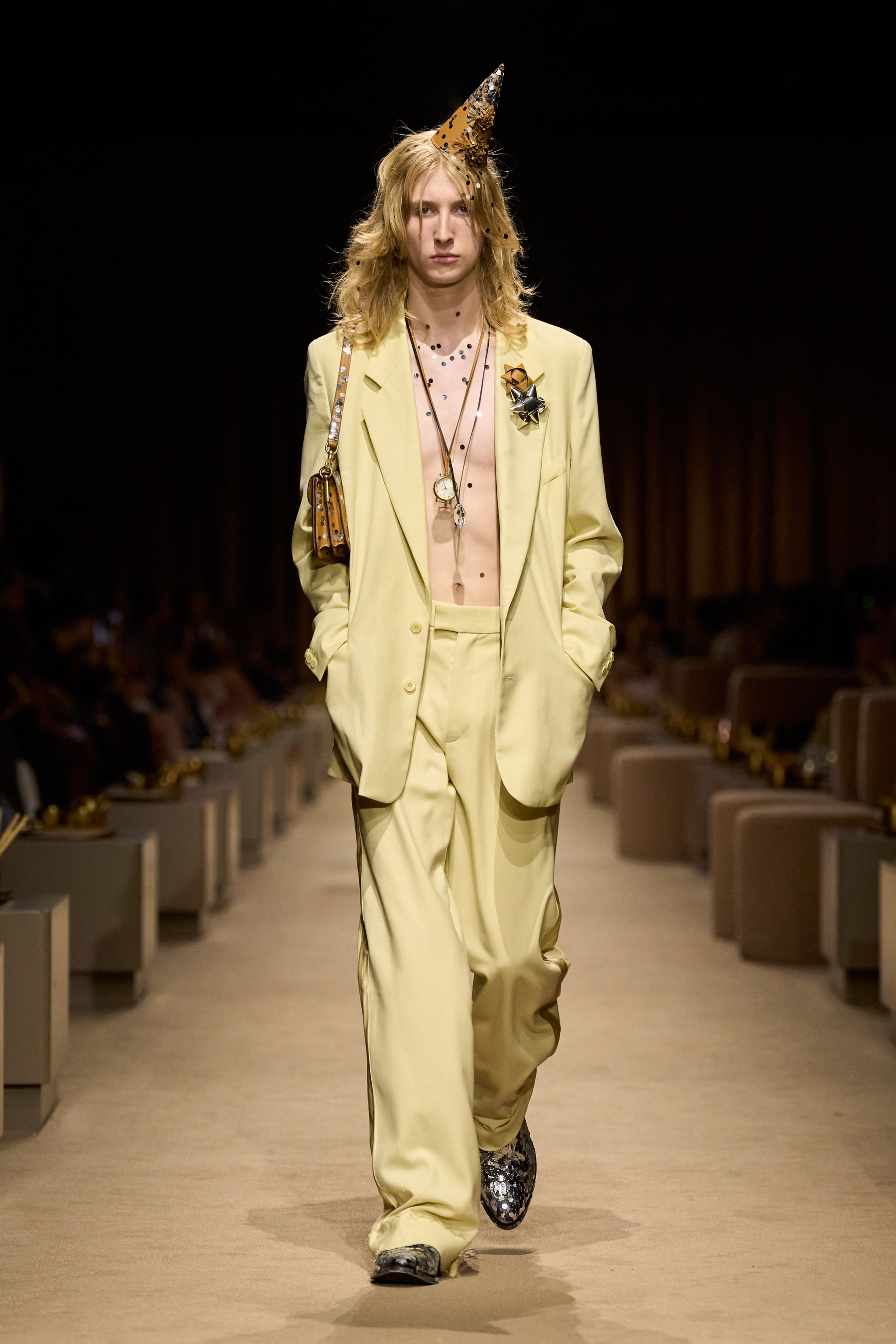 Coach Spring 2027 look 47: pale-yellow relaxed tailoring, layered necklaces, a gold bow brooch, metallic party hat and glittering shoes.