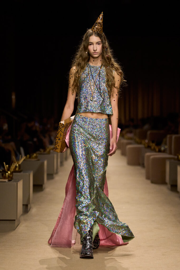 Coach Spring 2027 look 50: iridescent sequined top and long skirt with pink fabric panels, a pointed party hat and metallic boots on the runway.