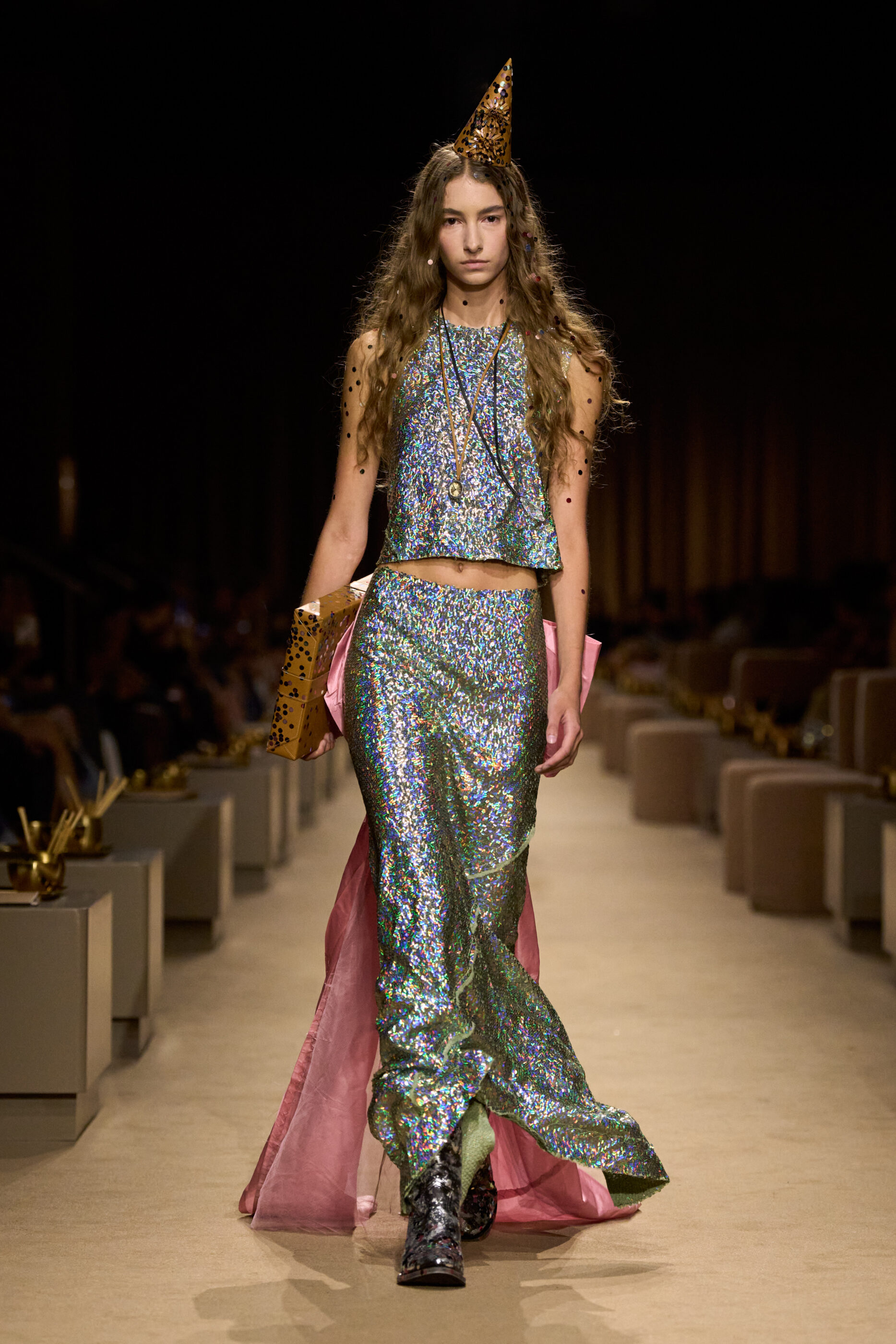 NYFW Spring 2027, Coach look 50: an iridescent sequined top and long skirt with pink fabric panels, a pointed party hat and metallic boots.