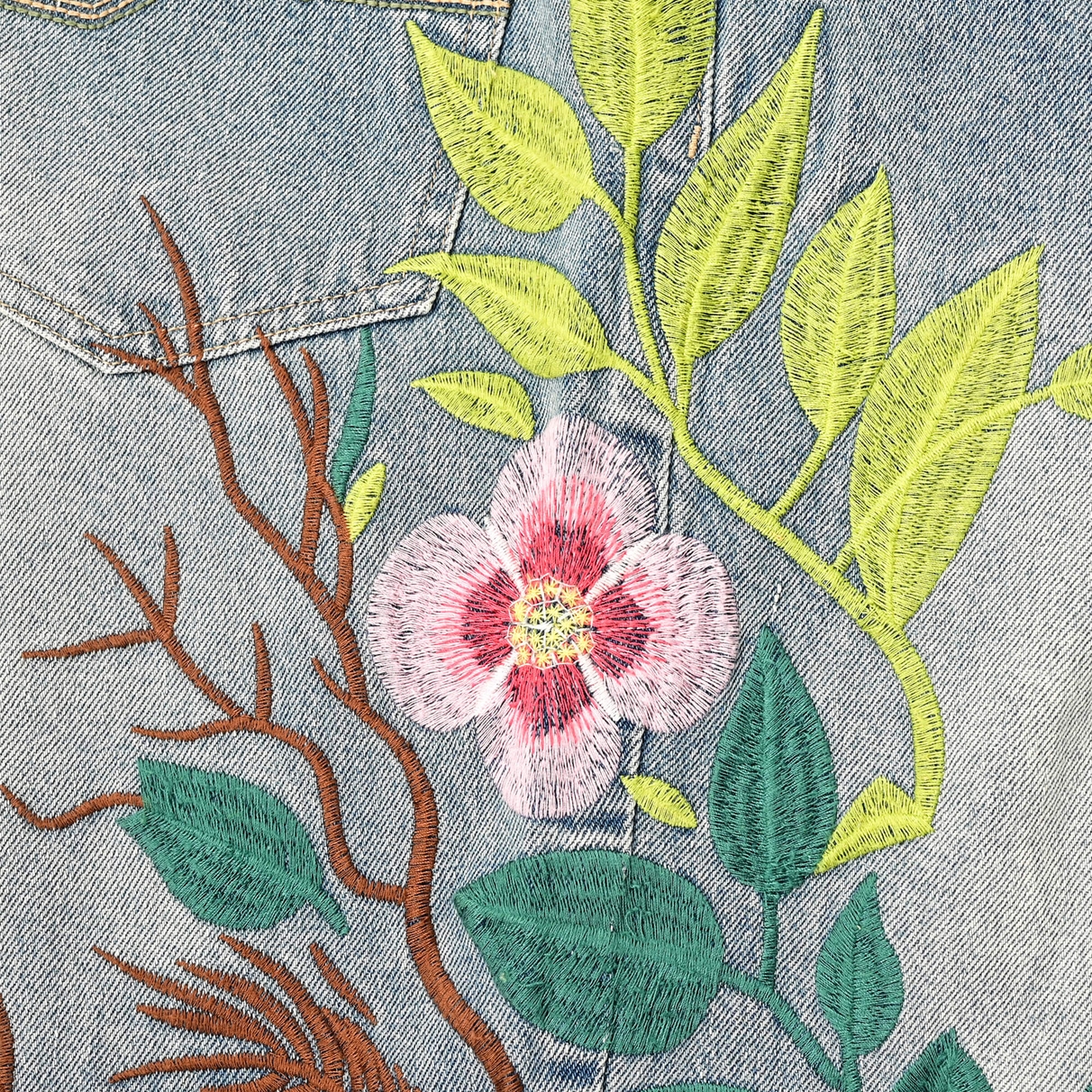 Close view of pink blossom and green leaf embroidery on the blue denim beside a pocket seam.