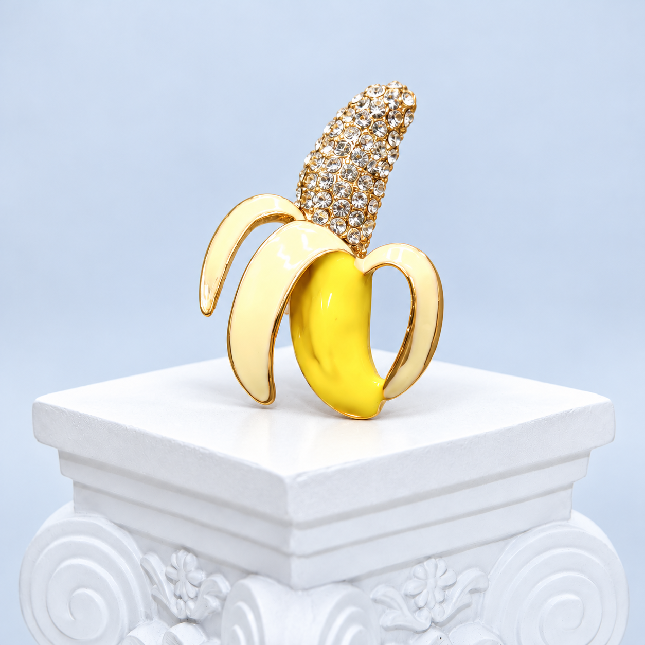 Front three-quarter view of the Mio Queena banana brooch with yellow and ivory enamel and a clear rhinestone top on a white pedestal