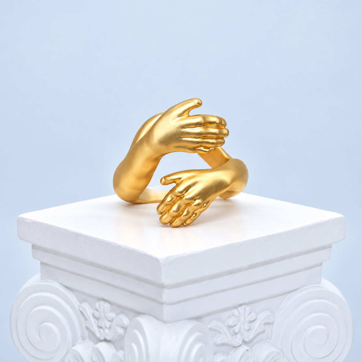 Front three-quarter view of the Mio Queena Embrace gold-plated copper ring with two sculpted hands on a white pedestal